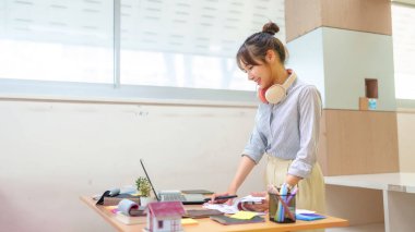 Asyalı genç kadın iç mimar dizüstü bilgisayar, kalem ve malzeme örnekleri kullanarak projeler üzerinde çalışıyor. Masasına bir ev modeliyle gülüyor yaratıcılık ve planlama sergiliyor.