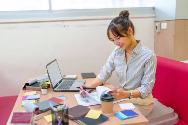 Asyalı genç bir yetişkin iç mimar dizayn planlarını gözden geçiriyor, dizüstü bilgisayar, renk örnekleri, tasarım kitapları ve modern bir ofiste bir kahve fincanı ile çevrili. İç tasarım kavramları için mükemmel.