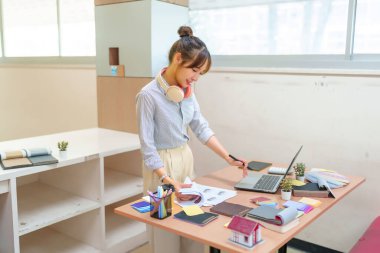 Asyalı genç kadın iç mimar, yaratıcı stüdyosunda malzeme örnekleri ev modelleri ve iç tasarım çizimleri ile çevrili bir dizüstü bilgisayarla masasında çalışıyor..