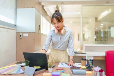 Genç bir Asyalı iç mimar renk örnekleri ve örneklerle dolu parlak bir ofisteki dizüstü bilgisayarda tasarımları analiz ediyor. İlham için tasarım sürecine ve çalışma alanına odaklanın.