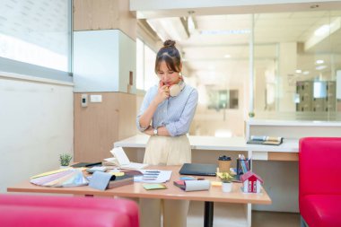 Genç bir Asyalı iç mimar masasında renk paletleri, kumaş örnekleri ve yaratıcı iç tasarım sürecini gösteren minyatür bir ev modeli ile çevrili yeni bir proje planlıyor..