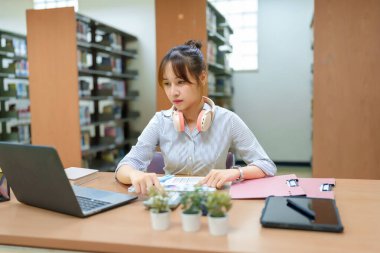 Asyalı genç bir öğrenci kütüphanede dizüstü bilgisayarla çalışıyor, belgeleri inceliyor. Eğitim, araştırma, akademik başarı ve sınav hazırlıklarına odaklanmış. Öğrenmeyi ve başarıyı geliştir.