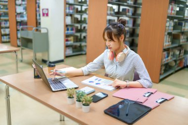Kütüphanede dizüstü bilgisayar kullanan genç Asyalı kadınlar. Başarı için eğitim ve akademik araştırmalara odaklanmak. Etrafı kitaplarla ve çalışma kitaplarıyla çevrili..