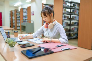 Kütüphanede dizüstü bilgisayar kullanan ve çizelgeleri analiz eden genç bir Asyalı kadın. Öğrenim, araştırma, öğrenim ve akademik başarı için veri analizine odaklanın.