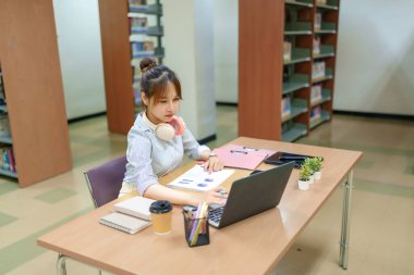 Kütüphanede dizüstü bilgisayarla okuyan genç bir Asyalı kadın resmi. Eğitim, araştırma, öğrenme ve öğrenim hayatına odaklan. Akademik başarı için kütüphane kaynaklarını ve ipuçlarını araştır.