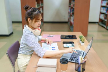 Görüntü, bir kütüphane ortamında dizüstü bilgisayarına odaklanmış genç bir Asyalı kadını gösteriyor. Çevrimiçi eğitim, üniversite hayatı, araştırma ve beceri geliştirme için ideal.