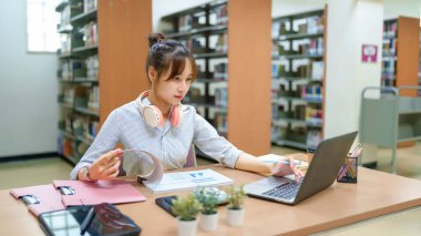 Genç bir Asyalı kadın kütüphanede dizüstü bilgisayar, grafik, ders kitabı ve kulaklık kullanarak ders çalışıyor. Çevrimiçi öğrenme, araştırma ve eğitime odaklanıyor. Bu kütüphane sessiz bir öğrenme ortamı sağlar..