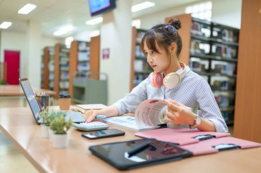 Genç bir Asyalı kadın kütüphanede okuyor, bilgisayarla, hesap makinesiyle ve tabletle finansal tabloları inceliyor. Kahve fincanı ve kitaplıklar yakınlarda. Eğitim, finans ve eğitim kavramları için mükemmel..