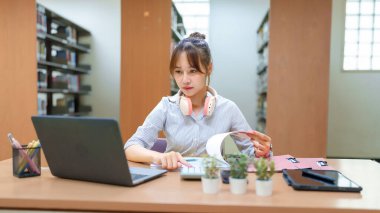 Asyalı genç bir kadın kütüphanede dizüstü bilgisayarı, hesap makinesi ve kitabıyla ders çalışıyor. Akademik araştırma ve sınavlara hazırlanırken, eğitimsel başarıyı somutlaştırıyor. Bu resim öğrenmeye adanmışlığı gösterir.