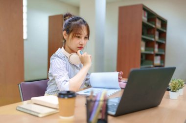 Kütüphane ortamında okuyan genç bir Asyalı kadın resmi. Dizüstü bilgisayar kullanıyor, belgeleri inceliyor ve kulaklıkları var. Eğitim, akademik araştırma ve öğrenim hayatı için ideal.