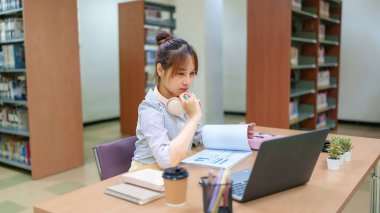 Genç Asyalı kadın kütüphanede dizüstü bilgisayarla veri tabloları üzerinde çalışıyor. Eğitim, araştırma ve finans grafiklerine odaklanmış. Üniversite, üniversite veya öğrenci öğrenimi görselleri için ideal.