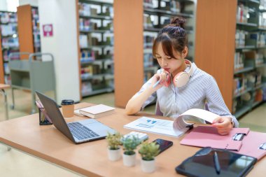 Kütüphanede okuyan genç bir Asyalı kadın resmi. Laptop kullanıyor, kulaklık takıyor ve verileri analiz ediyor. Bu resim eğitim, araştırma, akademik başarı ve kütüphane kaynaklarını temsil etmektedir..