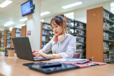 Kütüphanede dizüstü bilgisayar kullanan genç bir Asyalı kadın. Eğitim temalı fotoğraf eğitimi, öğrenimi, araştırması için. Laptop, kitaplar, kulaklıklar, tablet. Üniversite öğrencisi, eğitim kaynakları.