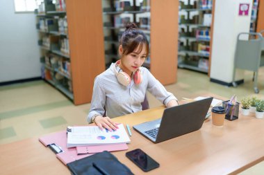 Genç bir Asyalı kadın arka planda kitaplarla birlikte bir kütüphanede dizüstü bilgisayarı üzerinde çalışıyor. Belgeleri inceliyor ve teknolojiyi öğrenmek ve araştırmak için kullanıyor. Eğitim ve öğrenci başarısına odaklanmış.