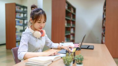 Kütüphanede ders çalışmaya odaklanmış genç bir Asyalı kadın görüntüsü. Bir dizüstü bilgisayar ve hesap makinesi kullanıyor. İnternetten öğrenme ya da araştırma öneriyor. Sahne eğitimi ve akademik başarıyı destekliyor.