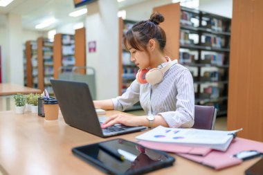 Genç Asyalı kadın kütüphanede okuyor, dizüstü bilgisayar kullanıyor. Eğitime, araştırmaya, çevrimiçi derslere, öğrenci başarısına odaklan. Kitaplıklar görünür. Akademik başarıyı ve öğrenim kaynaklarını destekle.