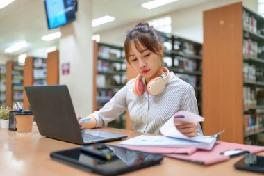 Kitaplarla çevrili bir kütüphanede dizüstü bilgisayarda çalışan genç Asyalı kadın. Belgeleri inceliyor, bir proje araştırıyor ve üniversitede eğitimini ilerletiyor. Eğitime ve öğrenmeye odaklanıyor.