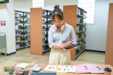 Kütüphane ortamında tabletine odaklanan genç bir Asyalı kadın. Eğitim içeriği, öğrenci hayatı, akademik çalışma, öğrenim teknolojisi ve kütüphane kaynakları için ideal.