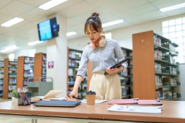 Genç Asyalı kadın kütüphane ortamında ders çalışıyor. Eğitim için laptopunu, tabletini ve diğer belgeleri kullanır. Bu, akademik bilgi arayışını araştırma ve başarı ile gösterir..