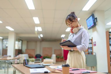 Kütüphane ortamında derslerine odaklanmış genç bir Asyalı kadının görüntüsü. O kulaklık takıyor ve araştırma için bir tablet kullanıyor. Eğitim içeriği, öğrenim hayatı veya akademik kaynaklar için mükemmel.