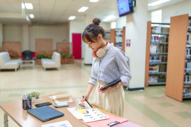 Genç bir Asyalı kadın kütüphanede veri tabloları üzerinde çalışıyor. Tablet kullanan bir üniversite öğrencisi, kitaplıklarla çevrili bir yüksek öğrenim ortamında belgeleri analiz ediyor. Kulaklık takıyor ve masadaki dosyalarına bakıyor..