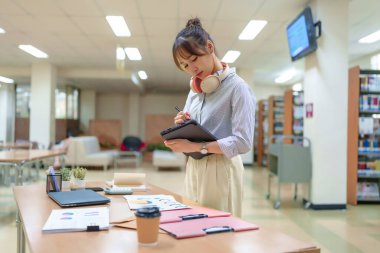 Yetişkin bir Asyalı kadın kütüphanede çalışıyor, tablet kullanıyor ve verileri analiz ediyor. O kulaklık takıyor. Araştırma ve eğitim kavramları tasvir edilir.