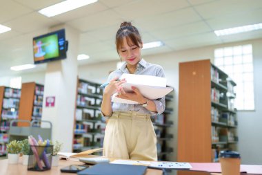 Kütüphanede kitap tutan ve not alan genç bir Asyalı kadın. Bir kütüphane ortamında eğitim, bilgi edinme, araştırma ve öğrenim materyallerine odaklanmış.