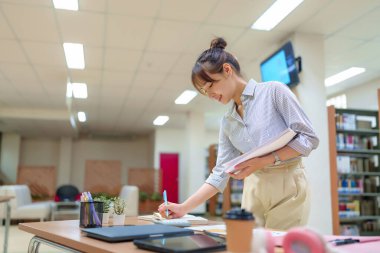 Genç Asyalı bir kadın kütüphane masasında kitap, dizüstü bilgisayarla çalışıyor ve eğitim için notlar alıyor. Akademik, bilgi, öğrenme, araştırma, başarı kavramı.