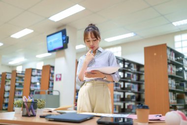 Asyalı genç bir kadının kütüphanede kitaplıklarla dolu defterini tutarken çekilmiş bir fotoğrafı. Bilgisayarı ve telefonu olan bir masanın yanında duruyor. Eğitim amacıyla araştırma ve öğrenim gördüğünü gösteriyor..