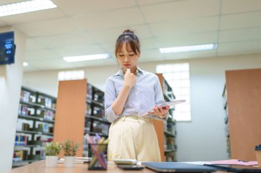 Genç bir Asyalı kadın kütüphanede çalışıyor, elinde belgeler var. Akademik bir proje için araştırma verilerini analiz ediyor. Eğitime, başarıya, konsantrasyona ve öğrenmeye odaklan.