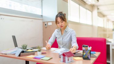 Genç bir Asyalı kadın iç mimar masasında dizüstü bilgisayar, boya örnekleri ve minyatür ev modeliyle planları inceliyor. Yaratıcı tasarıma odaklanmış durumda..