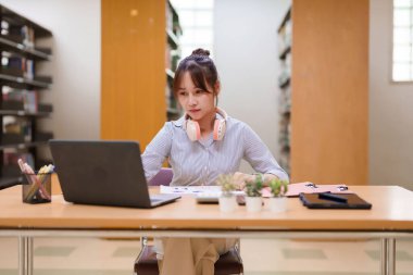 Kütüphanede dizüstü bilgisayarla okuyan Asyalı genç bir kadının resmi. Gösteri eğitimi, akademik araştırma, çevrimiçi öğrenme ve üniversite hayatı için mükemmel.