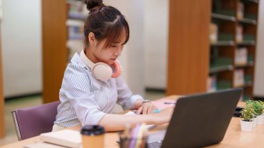 Genç Asyalı bir kadın dikkatle kütüphanede dizüstü bilgisayarıyla çalışıyor. Odaklanmış öğrenmenin, çevrimiçi eğitim kaynaklarının ve üniversite öğrencileri için akademik başarının önemini vurguluyor..