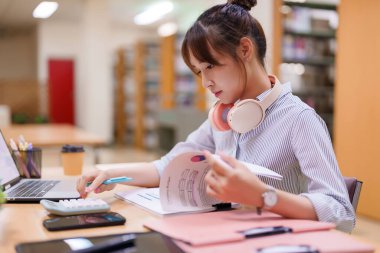 Genç bir Asyalı kadın kütüphane ortamında dizüstü bilgisayar, hesap makinesi ve yazılı raporlar kullanarak finansal verileri derinden inceliyor. Üniversite eğitimine, bütçe analizine ve araştırmaya odaklan.