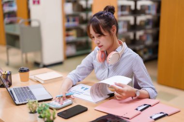 Genç Asyalı bir kadın kütüphanede okuyor. Bir dizüstü bilgisayar, hesap makinesi ve finansal tablolarla çalışıyor. Öğrenciler için eğitim, araştırma, akademik başarı ve kaynaklara odaklanın.