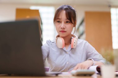Dizüstü bilgisayarına odaklanmış, kulaklık takan genç bir Asyalı kadın resmi. Bu, bir kütüphane ortamında modern eğitim, çevrimiçi öğrenme ve öğrenci hayatını tasvir etmektedir. Çalışmaya ve teknolojiye odaklan..