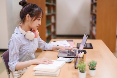 Genç Asyalı kadın kütüphanede dizüstü bilgisayar kullanıyor. Çevrimiçi çalışmalarına ve ödevlerine odaklanmış, akademik bir proje üzerinde çalışıyor ve üniversite ödevleri için araştırma yapıyor..