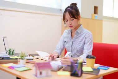 Asyalı yetişkin bir kadın dizüstü bilgisayarında iç dizayn okuyor. Dizüstü bilgisayarı, model evi, yapışkan notları ve kahvesi olan bir masada çalışıyor. İç mimari içeriği için mükemmel..