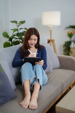 Bir Asyalı kadın (20 'li-30' lu yaşlarda), dijital sanat veya iş için stili olan bir tablet kullandığı için rahat bir şekilde koltuğunda oturuyor. Ev sahnesi dinleniyor.