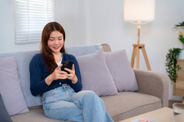 Neşeli bir Asyalı kadın (20 'li 30' lu yaşlarda) rahat bir kanepede, akıllı telefonuna göz atmaktan zevk alır. Modern ve rahat bir oturma odası. Yaşam tarzı ve teknoloji temaları için mükemmel.