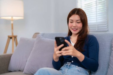 Genç bir Asyalı kadın (20-30 'lu yaşlarda) oturma odasında bir kanepede oturuyor. Gülümsüyor ve akıllı telefonunu kullanıyor. Ev rahatlama kavramı.