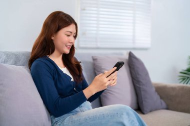 Genç bir Asyalı kadın (20-30 yaşlarında) akıllı telefonunu kullanarak bir kanepede rahatça dinleniyor. Hırka ve kot giyiyor, keyfine bakıyor..