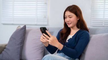 Genç bir Asyalı kadın (20-30 'lu yaşlarda) evdeki bir kanepede rahat bir şekilde akıllı telefonunu kullanıyor. Gülümsüyor, rahat giyiniyor ve sohbet ederken ya da gezinirken rahatlamış görünüyor..