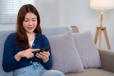 20 'li yaşlarda Asyalı bir kadın evinde rahat bir kanepede dinlenirken akıllı telefonunu rastgele kullanıyor. Mavi bir hırka ve kot pantolon giyiyor, boş zamanlarının tadını çıkarıyor..