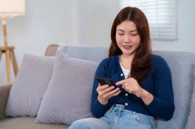 Genç bir Asyalı kadın (20-30 'lu yaşlarda) akıllı telefonunu kullanırken evdeki bir kanepede dinleniyor. Günlük giysiler giyiyor ve boş zamanlarının keyfini çıkarıyor gibi görünüyor..