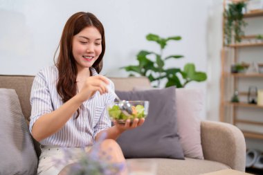 20 'li yaşlarda genç bir kadın kanepede taze salata yerken gülümsüyor. Sağlıklı beslenme, sağlıklı yaşam tarzı ve genç yetişkinler için dengeli beslenme kavramı.