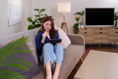 Evde bir kanepede tablet ve stil kullanan genç bir kadının görüntüsü. Uzaktan çalışma, çevrimiçi öğrenme ya da boş zaman aktivitelerini resmetmek için mükemmel. Modern iç mekan, rahat ve şık..