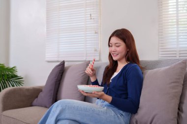 Genç bir kadın (20-30 'lu yaşlarda) kanepeye uzanır ve bir tabak atıştırmalık ekmeğin keyfini çıkarır. Parlak, rahat bir ev ortamında gündelik hayat sahnesi. Sağlıklı atıştırma ve dikkatli yemek temaları için mükemmel..