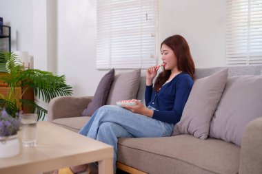 Genç bir Asyalı kadın (20-30 'lu yaşlarda) oturma odasındaki bir kanepede rahatça oturup tatlı bir atıştırmalığın tadını çıkarıyor. Bu rahat ve rahat sahne, evdeki basit bir zevk anını yakalıyor..