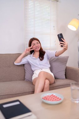20 'li yaşlarındaki neşeli genç bir kadın evdeki bir kanepede dinlenirken telefonuyla selfie çekiyor. Parmaklarıyla barış işareti yapıyor. Sıradan moda ve rahat atmosfer.